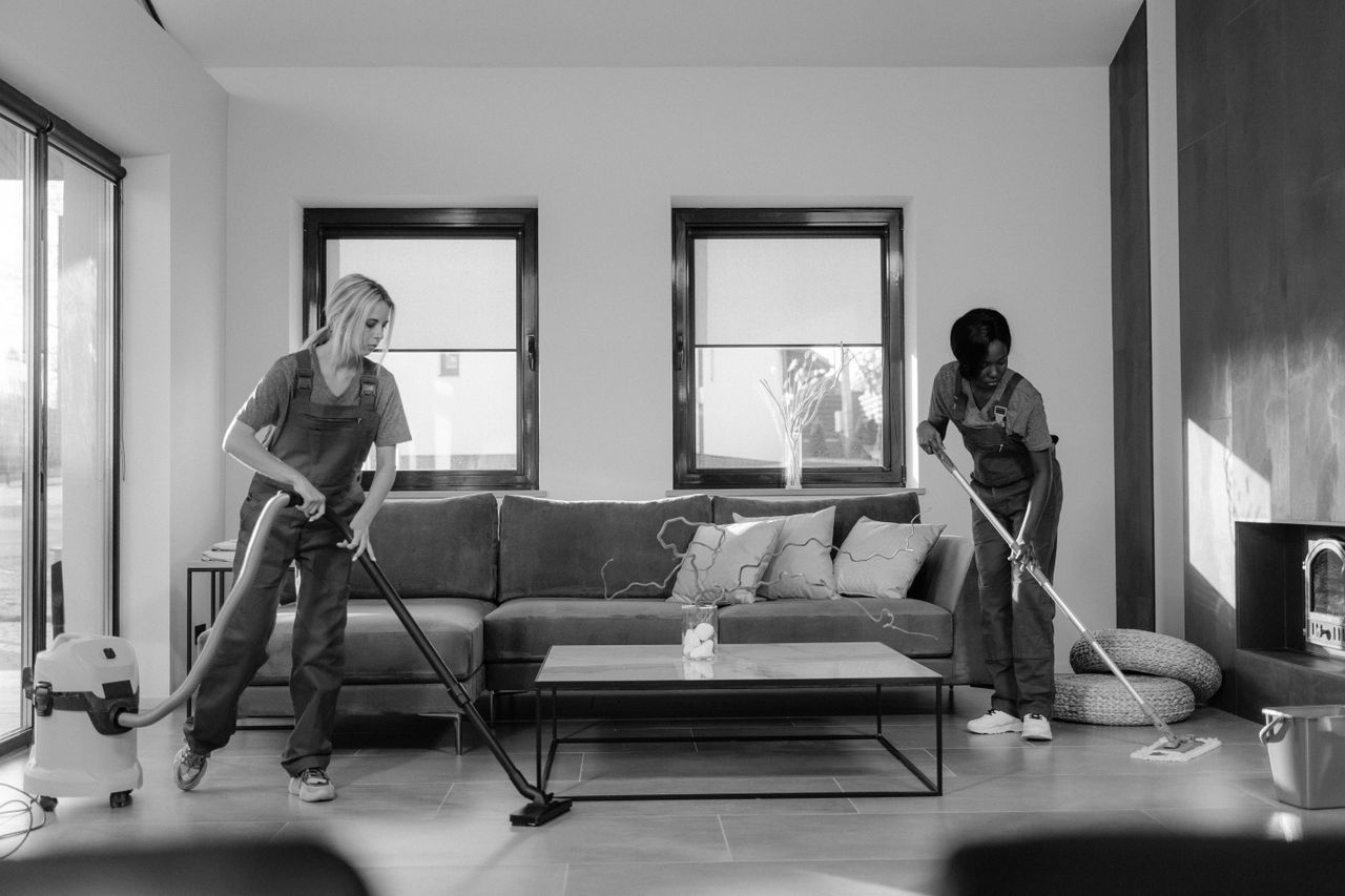 portfolio-06 Two women in work clothes cleaning a modern living room using a vacuum and mop.